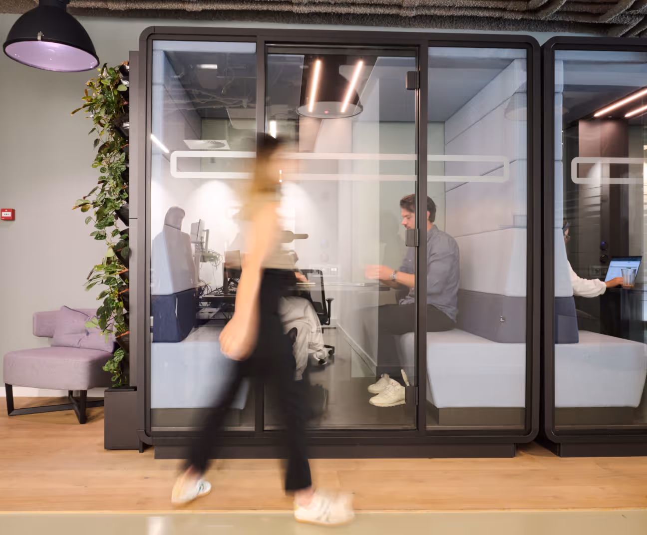 Modern office with a woman walking past glass-walled meeting pods where people work on laptops.