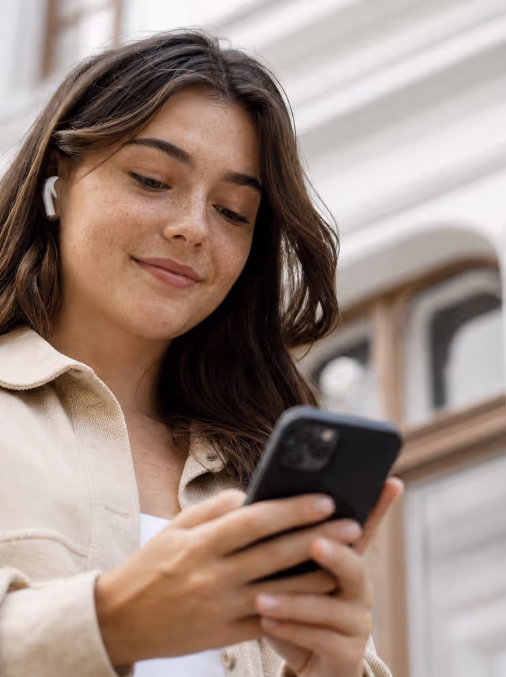 Smiling young woman wearing wireless earbuds looking at her smartphone outdoors.