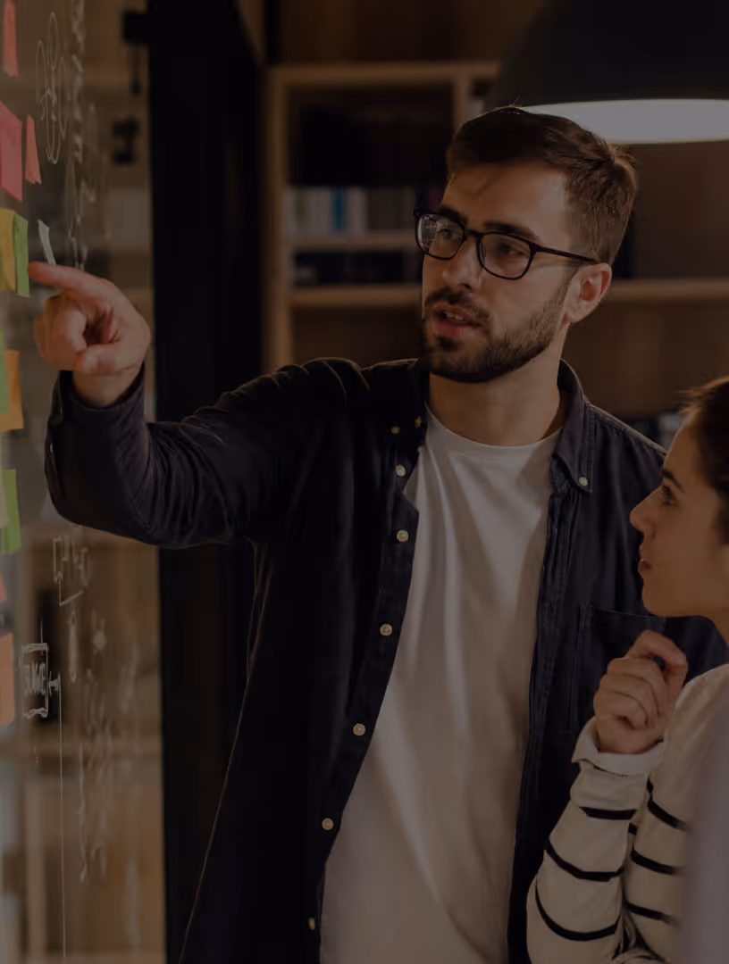 Man in glasses points at sticky notes on a glass wall while woman listens thoughtfully.