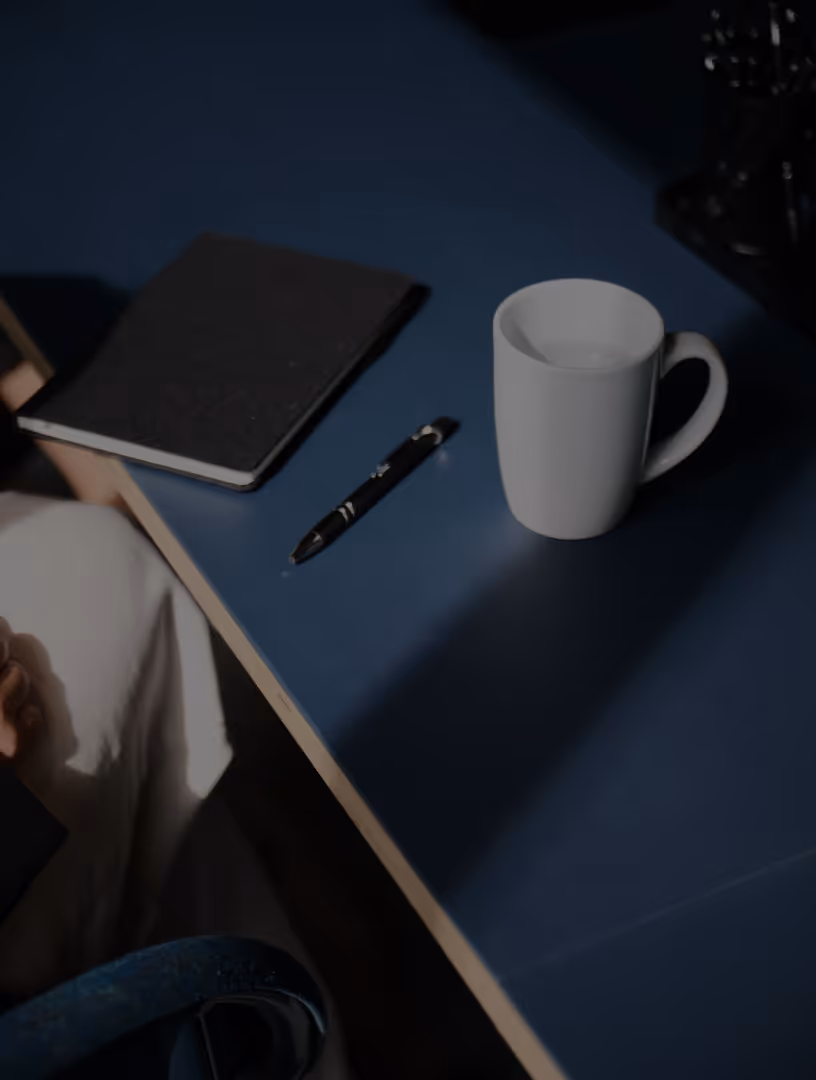 White coffee mug, black pen, and closed notebook arranged on a blue desk surface.