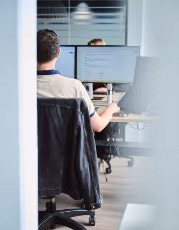 Man in beige shirt working on dual monitors in a modern office setting.