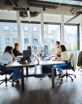 Four people working on computers together at a bright office with large windows.