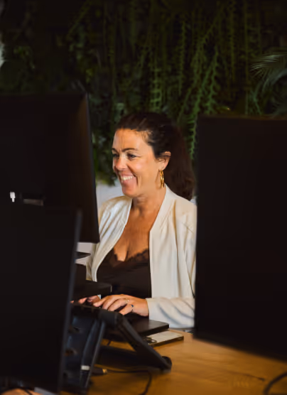 Smiling woman wearing white blazer working at a computer desk with green plants in the background.