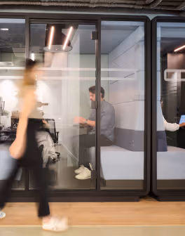 Blurred person walking past glass office wall where two people work inside.