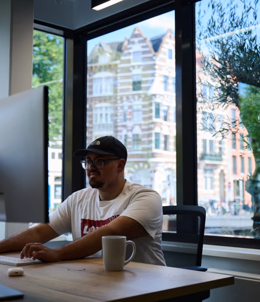 Person wearing glasses and cap working on computer at desk with mug and window showing building outside.