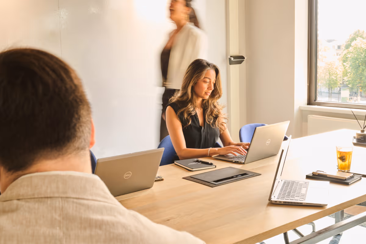 Woman typing on a laptop at a conference table with two other laptops and a blurred person walking by.