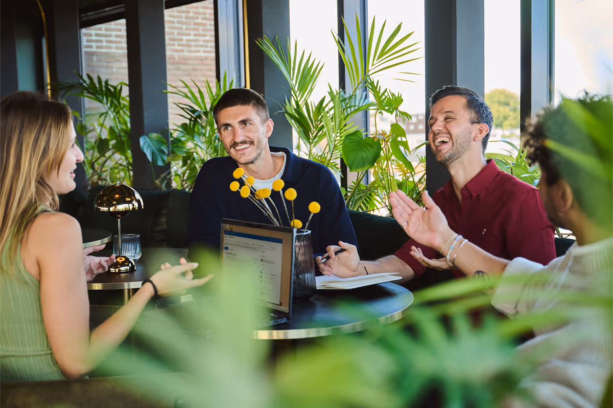 Four young adults sitting around a table in a plant-filled room, speaking and laughing together.