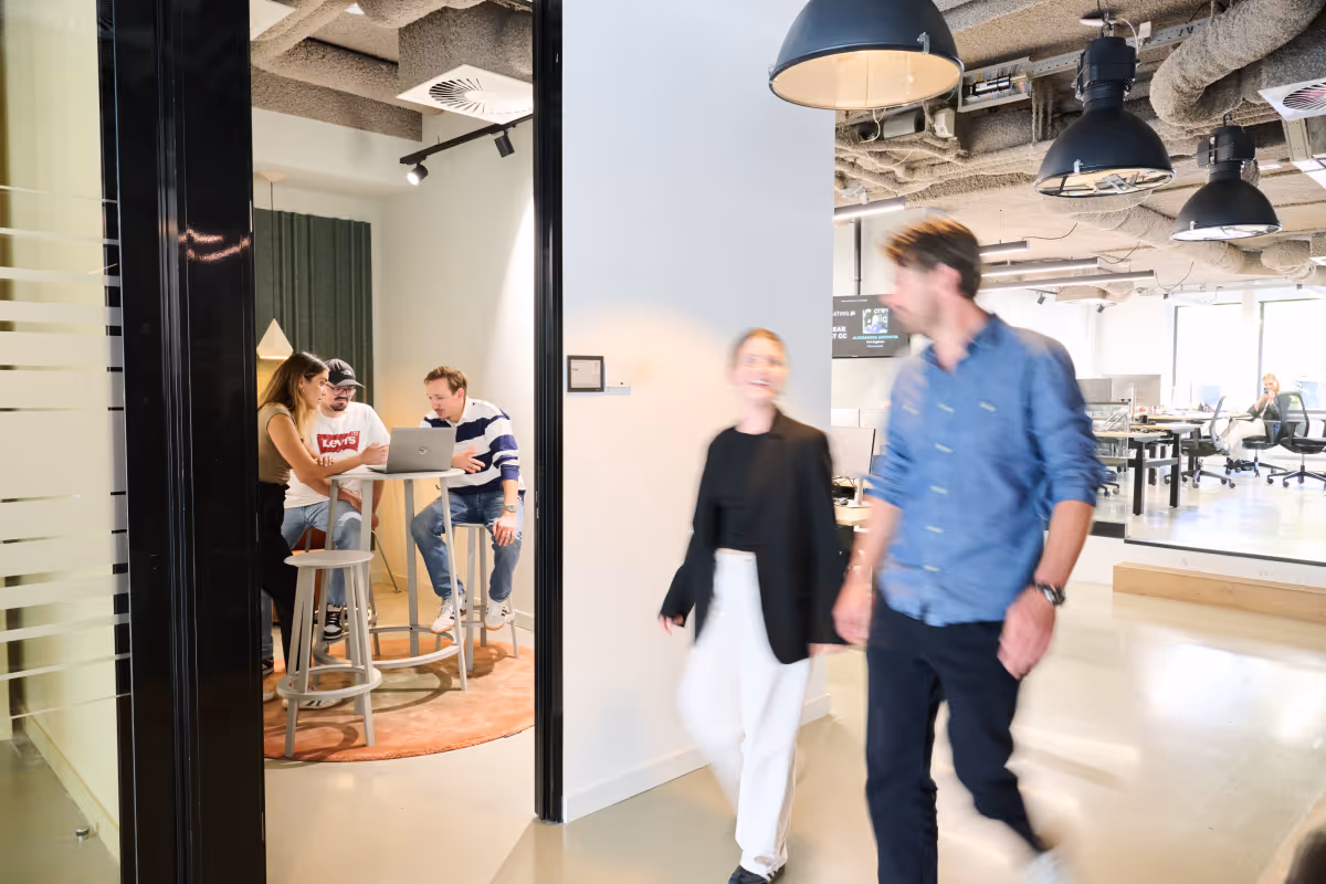 Three people work at a laptop in a small meeting room while two others walk by in an open office.