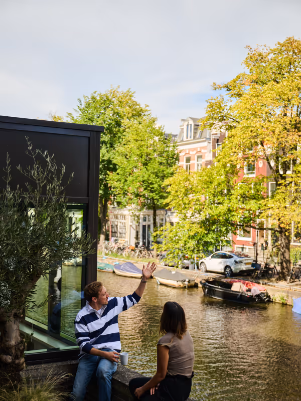 Two people sitting by a canal in a city with trees, boats, and buildings in the background.