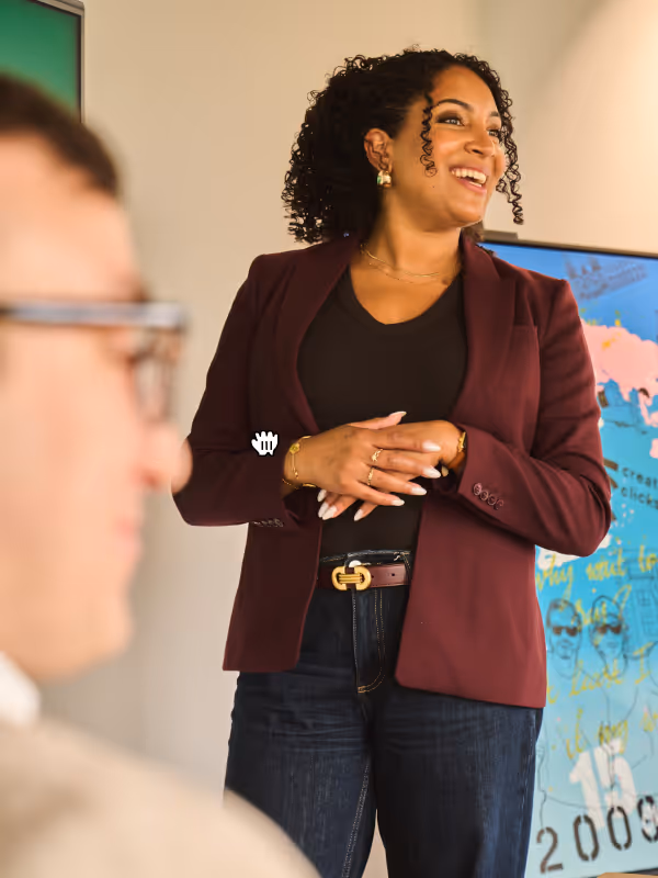 Smiling woman in a burgundy blazer talking in an office with a blurred person in foreground.