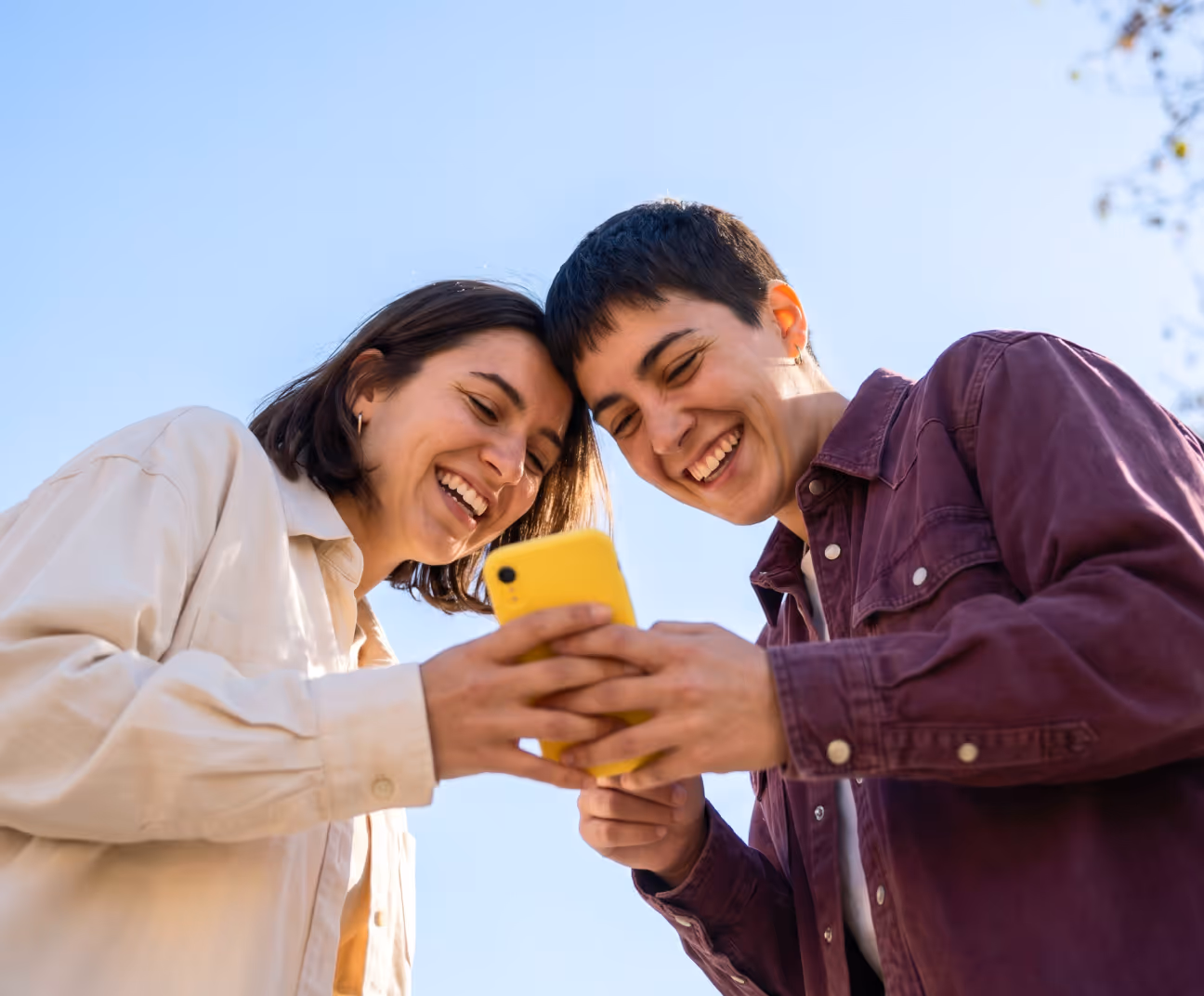 Two young people smiling and looking at a yellow smartphone outdoors under a clear blue sky.