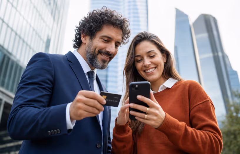 Smiling man with credit card and woman with smartphone outside modern skyscrapers.