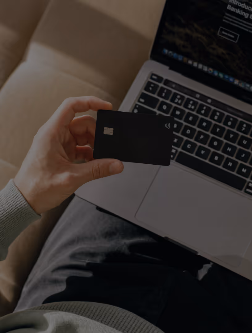 Person holding a black contactless credit card near a laptop keyboard on their lap.