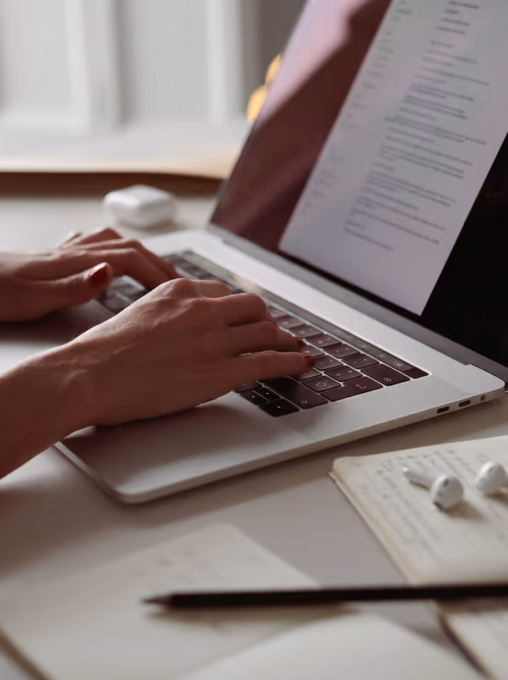 Hands typing on a laptop keyboard with a blurred screen and notebook with earbuds nearby.