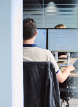 Man working on dual monitors in an office with glass walls and blinds.