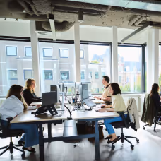 People working on computers at desks inside an office with large windows and natural light.