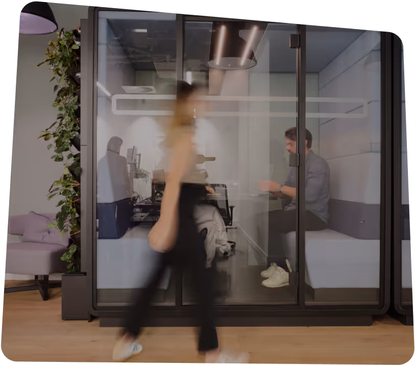 Man sitting in glass office booth working on laptop while woman walks past in modern workspace.