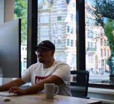 Man in glasses and cap working on a laptop at a table by a large window with a city view.