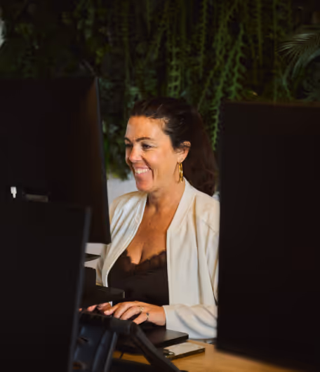 Smiling woman with dark hair working on a computer in an office with green plants in the background.