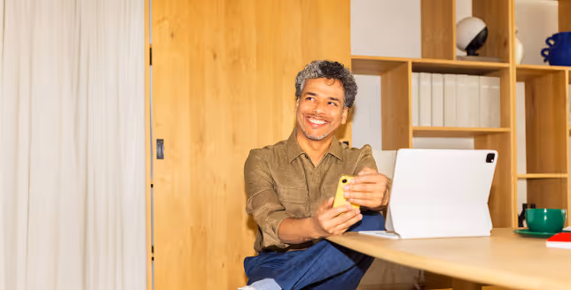 Man smiling and holding a yellow phone, sitting at a table with a tablet and cup in a modern room.