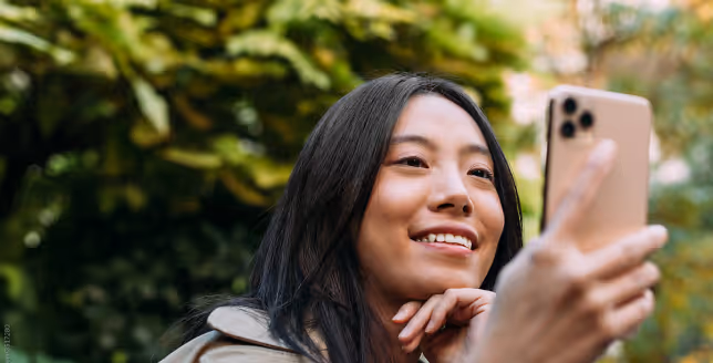 Young woman with long dark hair smiling outdoors while holding a gold smartphone for a selfie.