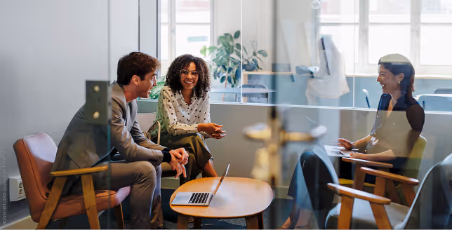 Three diverse colleagues sitting and chatting in an office with a laptop on a wooden table.