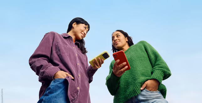 Two women smiling and looking at each other while holding smartphones against a clear sky.