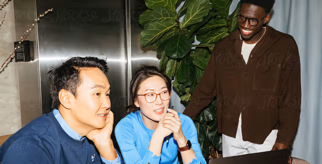 Three diverse coworkers smiling and discussing work around a laptop in an office setting.