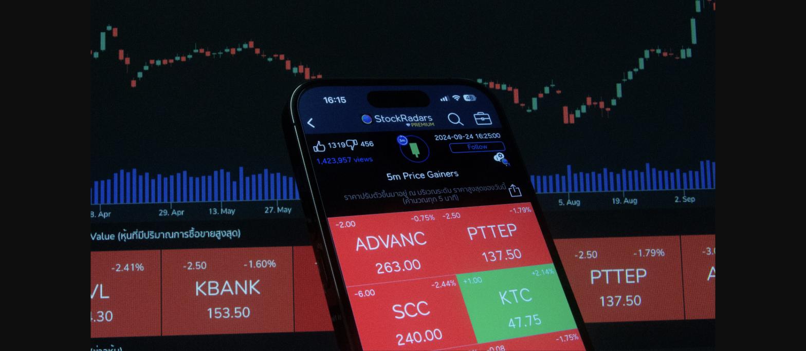 Smartphone showing stock market price gainers amidst background of stock charts and volume bars.