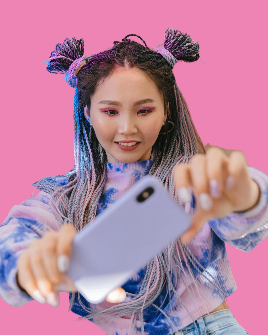 Young woman with braided hair styled in two buns wearing a blue tie-dye sweater, holding a smartphone and taking a selfie against a pink background.