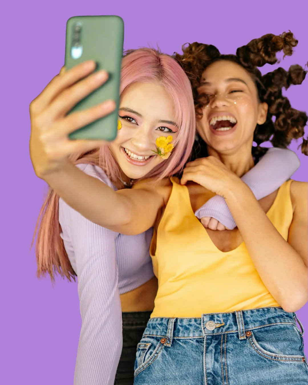 Two young women smiling and taking a selfie together against a purple background, one with pink hair and yellow flower decorations on her face, the other with curly hair and wearing a yellow top.