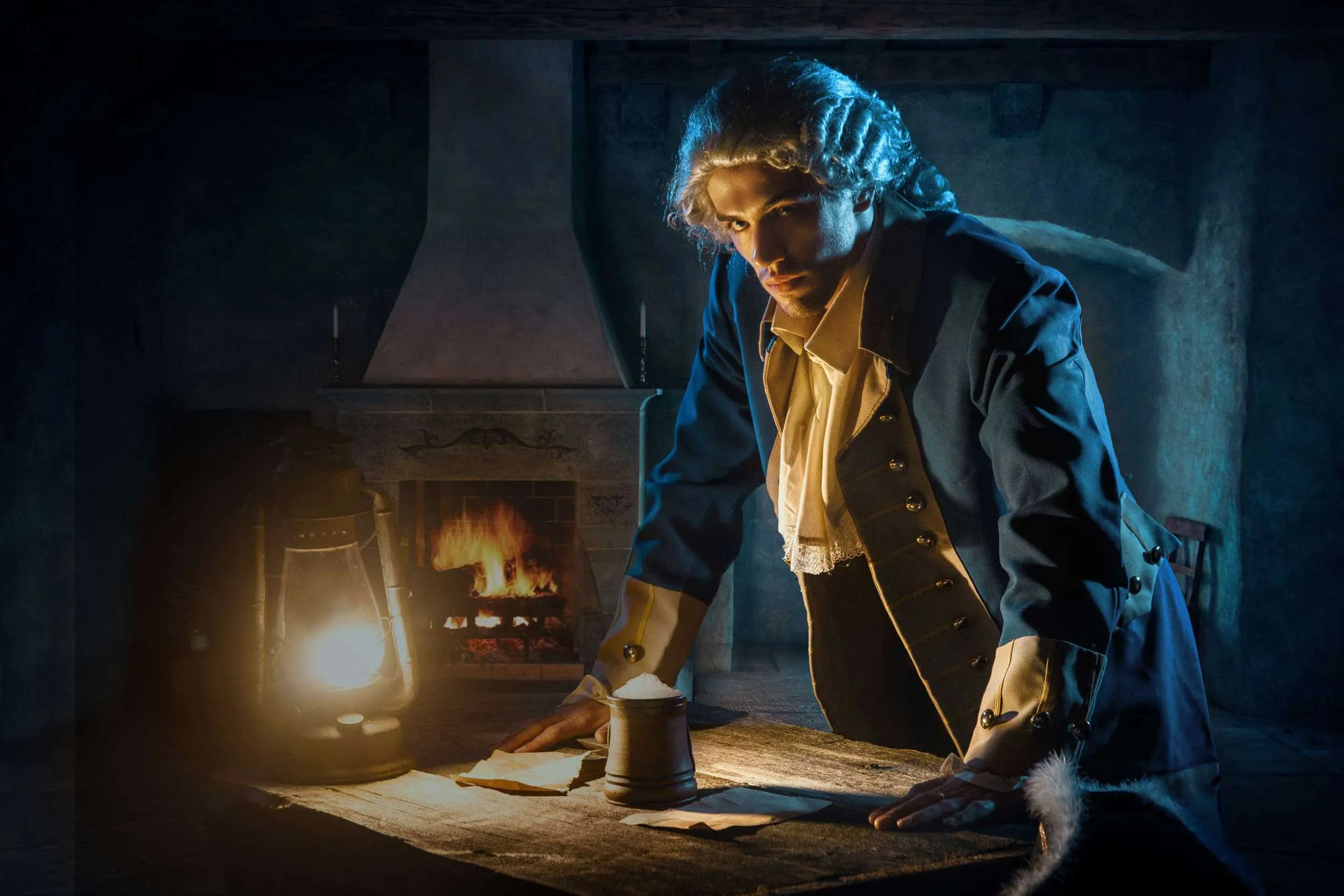 Man in historische kleding leunt op tafel met kaarslicht en open haard op achtergrond.