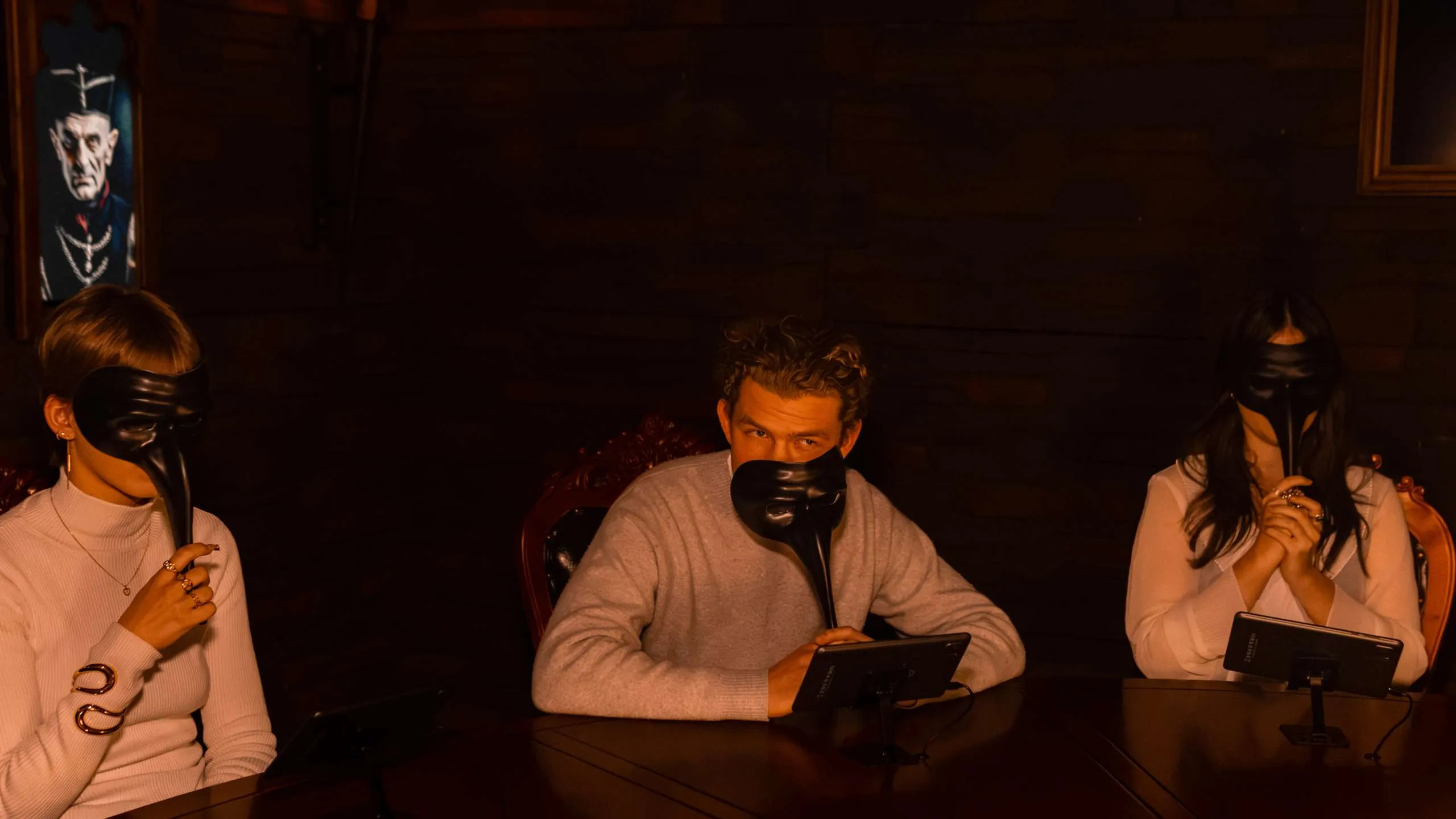 Drie mensen zitten aan een tafel in een donkere kamer met lange zwarte maskers op hun gezichten.