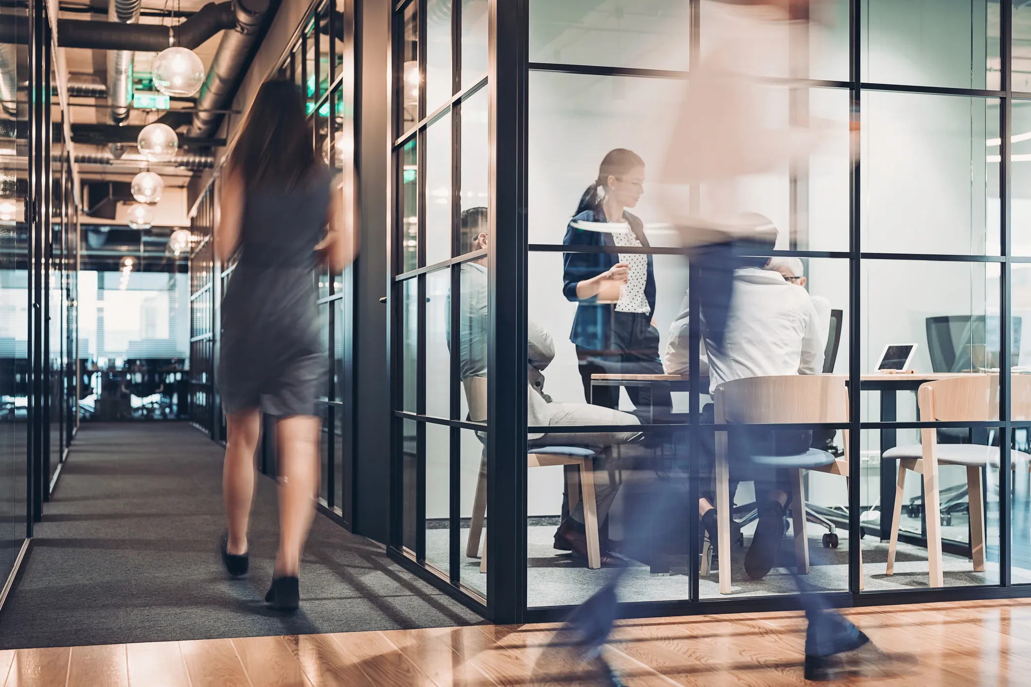 Blurred people walking past a glass-walled office with colleagues having a meeting inside.