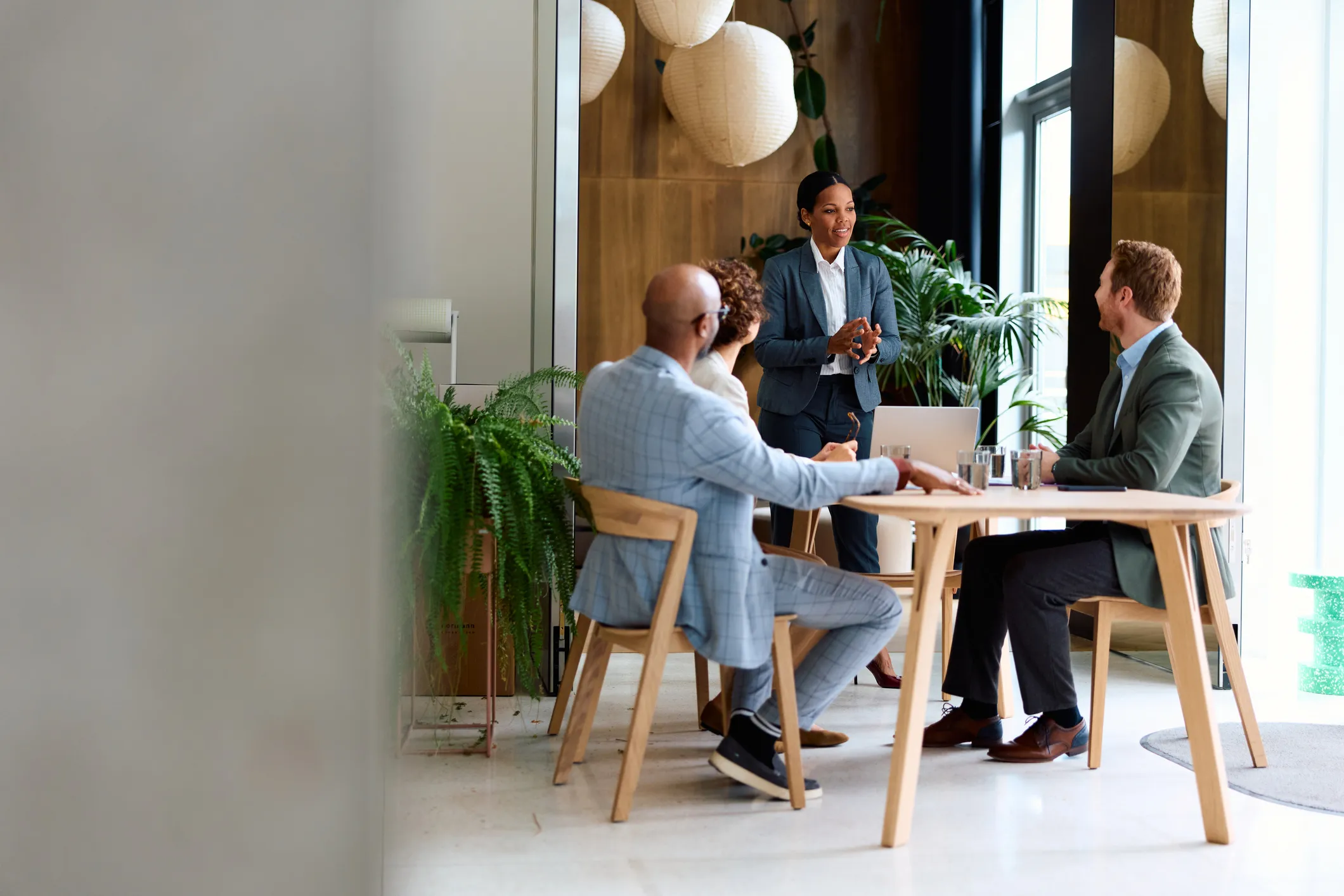 Businesswoman standing and speaking to three seated colleagues in a modern office with plants.