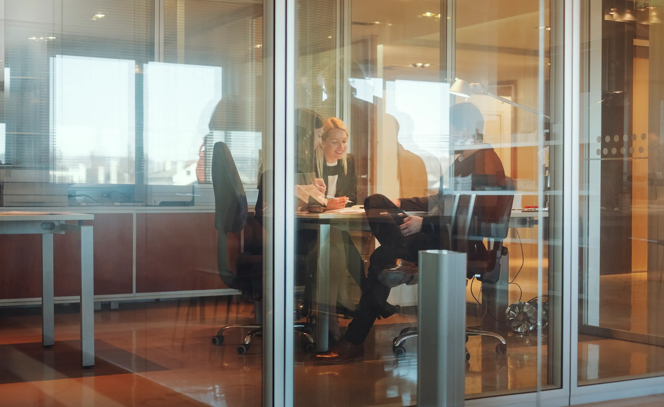 Three professionals having a meeting inside a glass-walled office with city view windows.