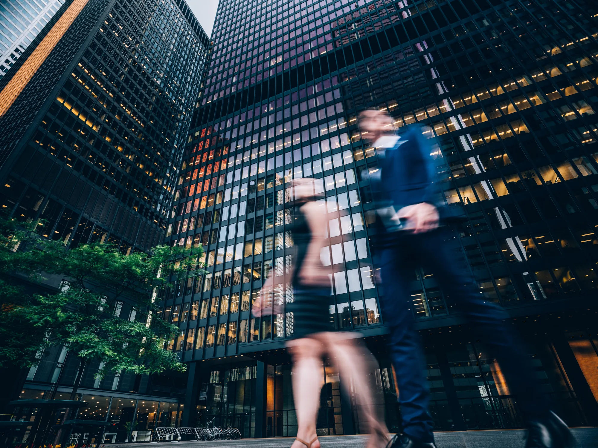 Blurred motion of two business people walking past tall glass office buildings in a city.