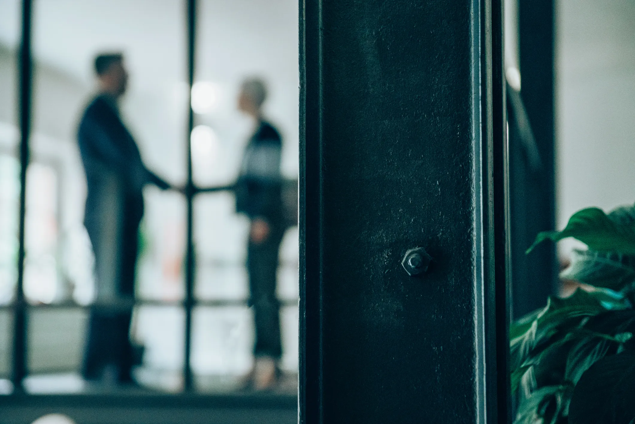 Blurred image of two people shaking hands in an office with glass walls and plants.