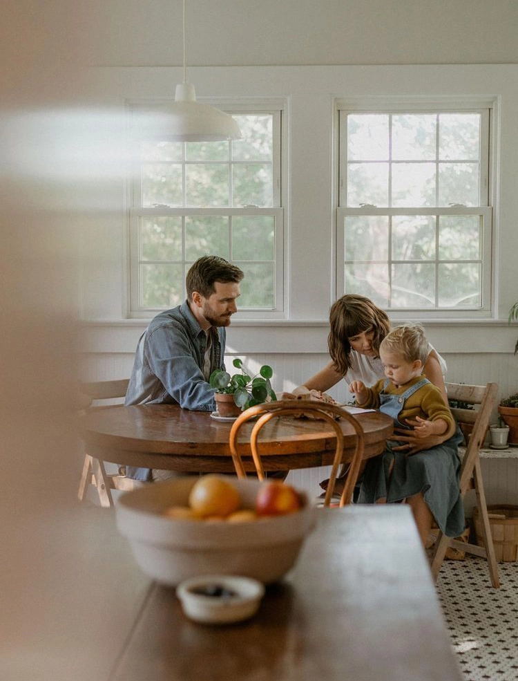 Man and woman with toddler sitting at a round wooden table near two large windows in a cozy kitchen.
