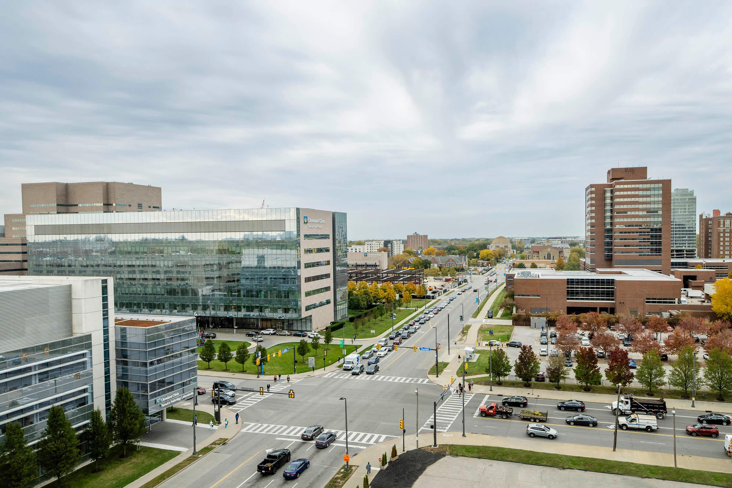 City intersection with traffic, modern office buildings, and trees under a cloudy sky.