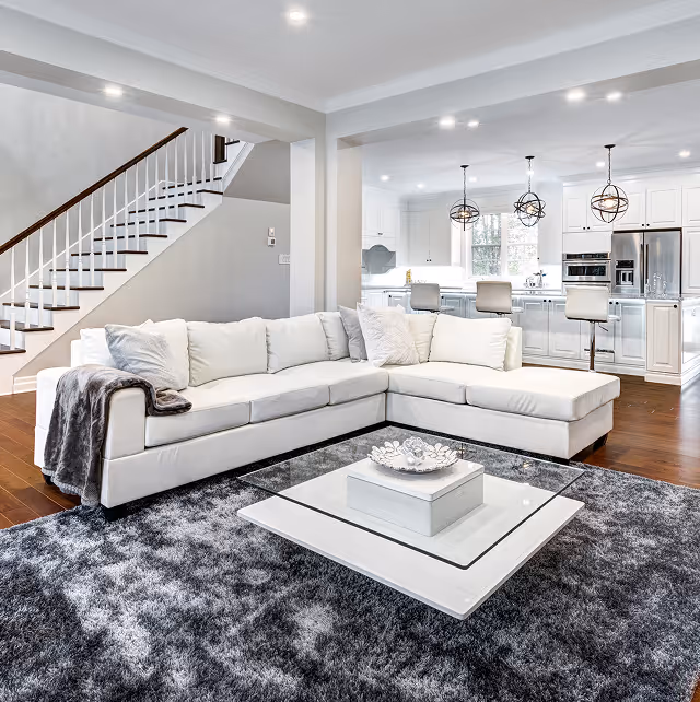 Bright living room with white sectional sofa, glass coffee table, gray rug, and open kitchen in background.