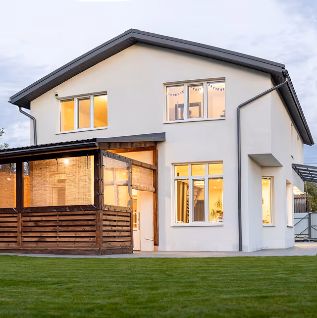 Modern two-story house with white walls, lit windows, wooden porch, and green lawn in front.