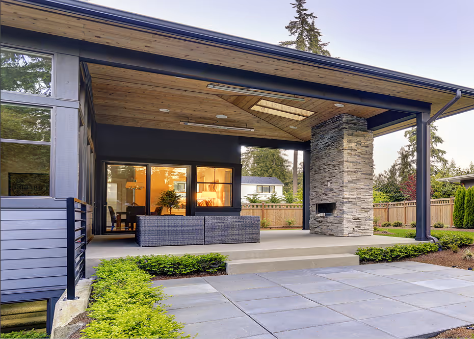 Covered patio with stone fireplace, wicker furniture, and wooden ceiling in a backyard with plants.