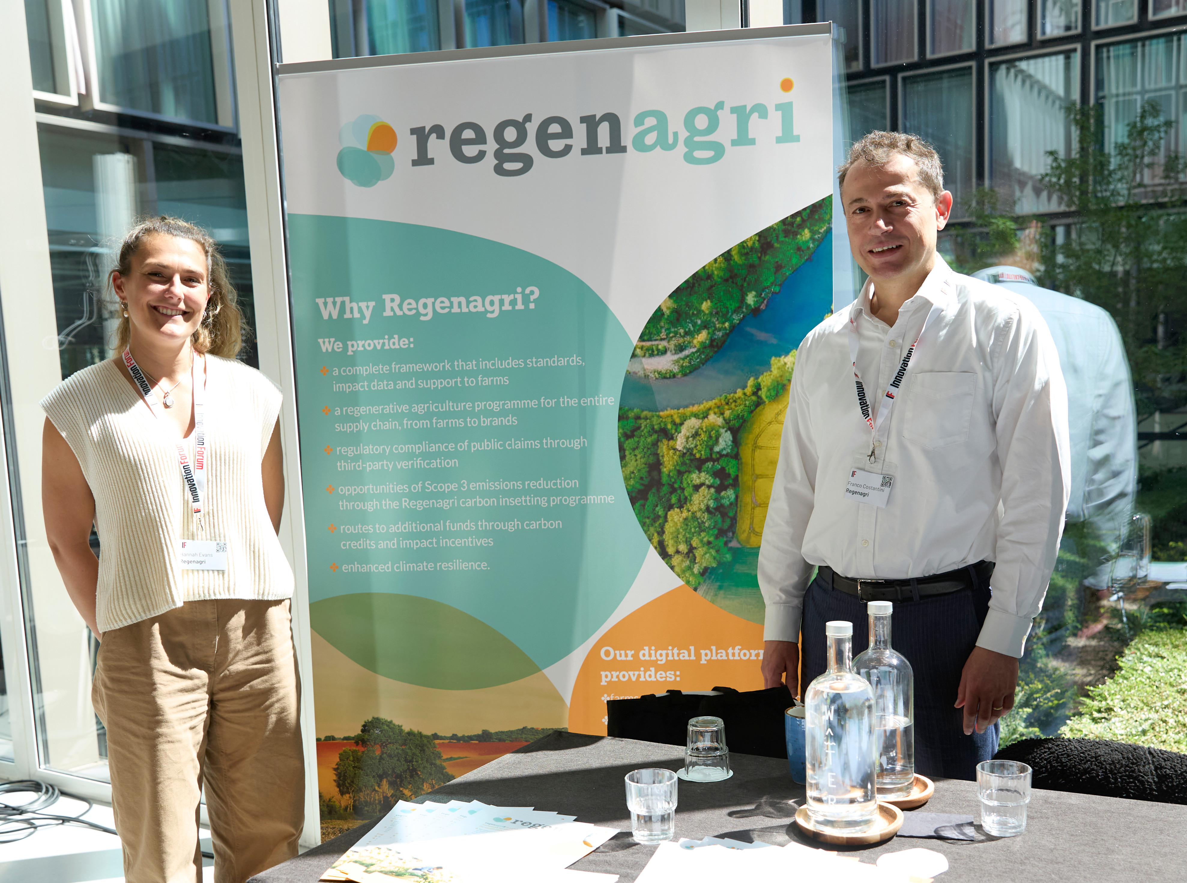 Two Regenagri representatives standing beside a Why Regenagri? pull-up banner at an indoor event stand