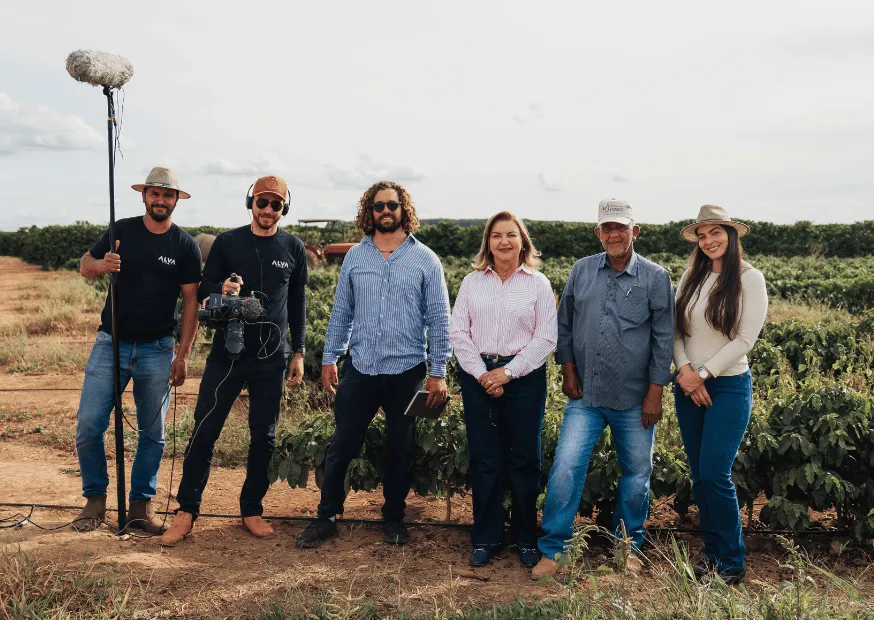 A two-person film crew with boom mic and camera rig standing with four farm hosts beside plantation rows