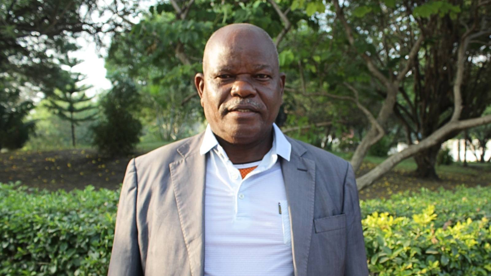 Mr John Kabole of Alliance Ginneries, in a grey jacket and white polo shirt, standing among trees and hedges