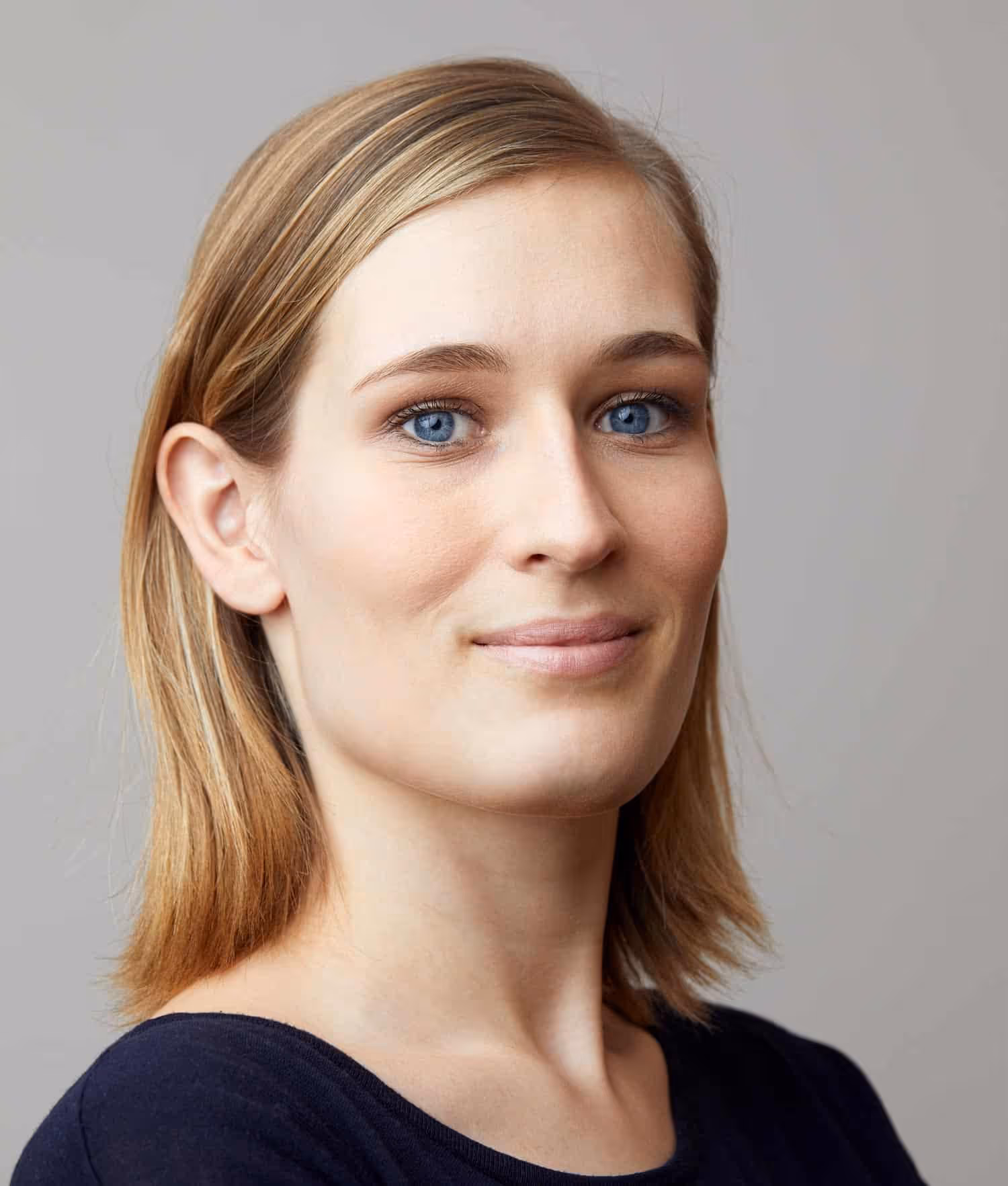Portrait of a young woman with blonde hair and blue eyes wearing a dark top against a gray background.