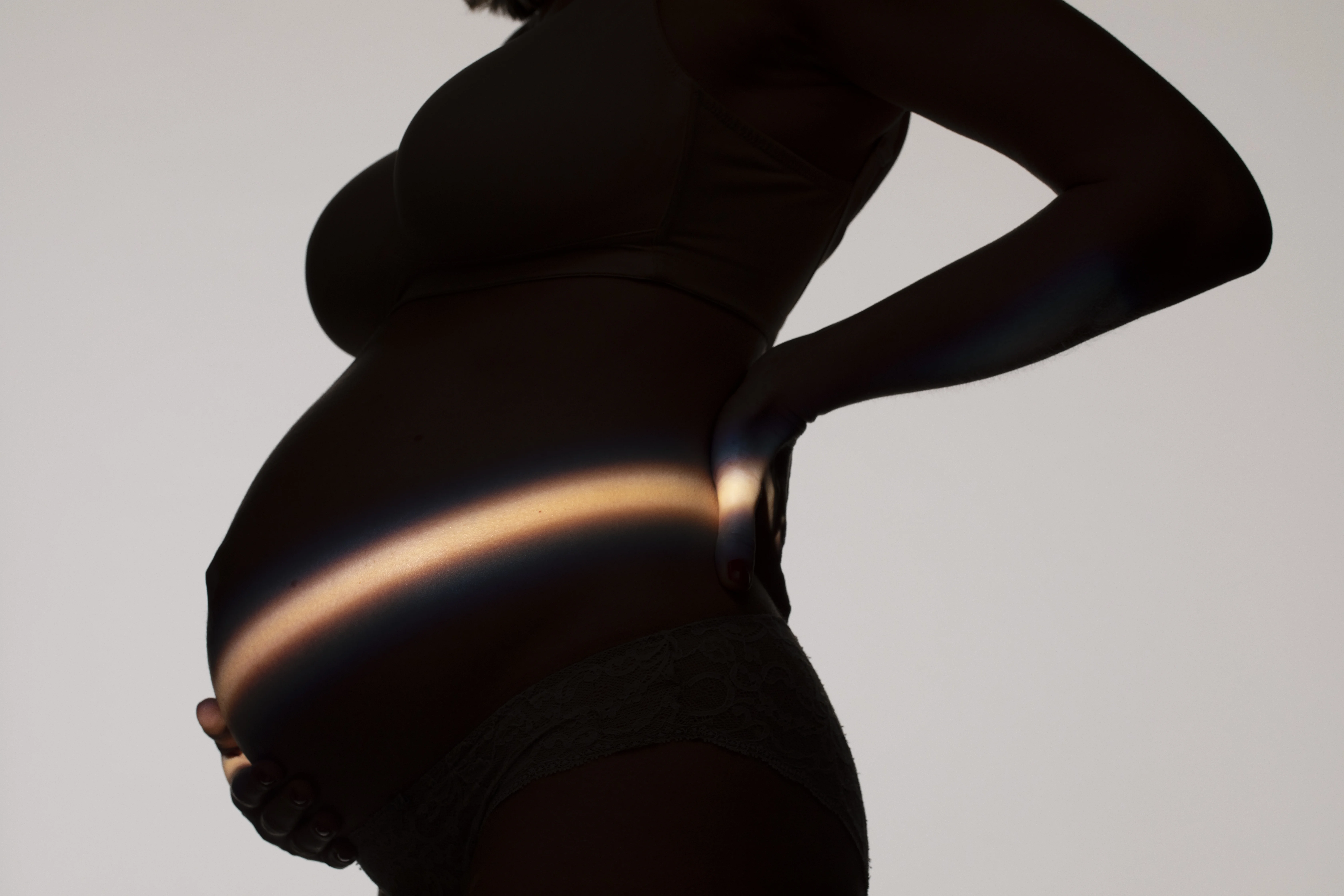 Silhouette einer schwangeren Frau im Profil vor einem Mommy Makeover, kunstvoll inszeniert mit einem Lichtstreifen auf dem Babybauch.