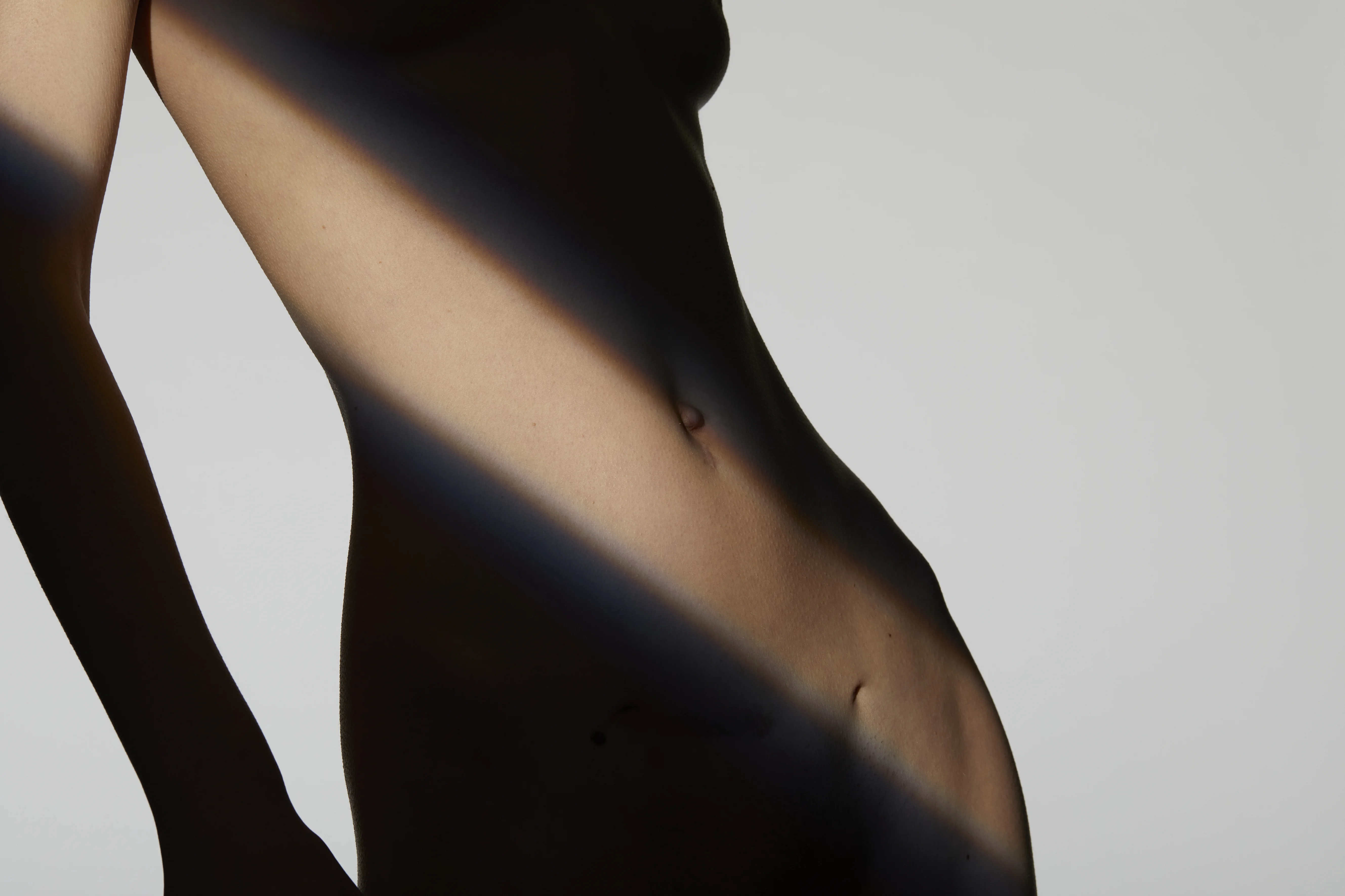 Kunstvolle Nahaufnahme eines weiblichen Torsos und Bauchs mit einer feinen Linie im Unterbauchbereich, akzentuiert durch ein Licht- und Schattenspiel – passend zum Thema Narbenbehandlung und Narbenkorrektur.