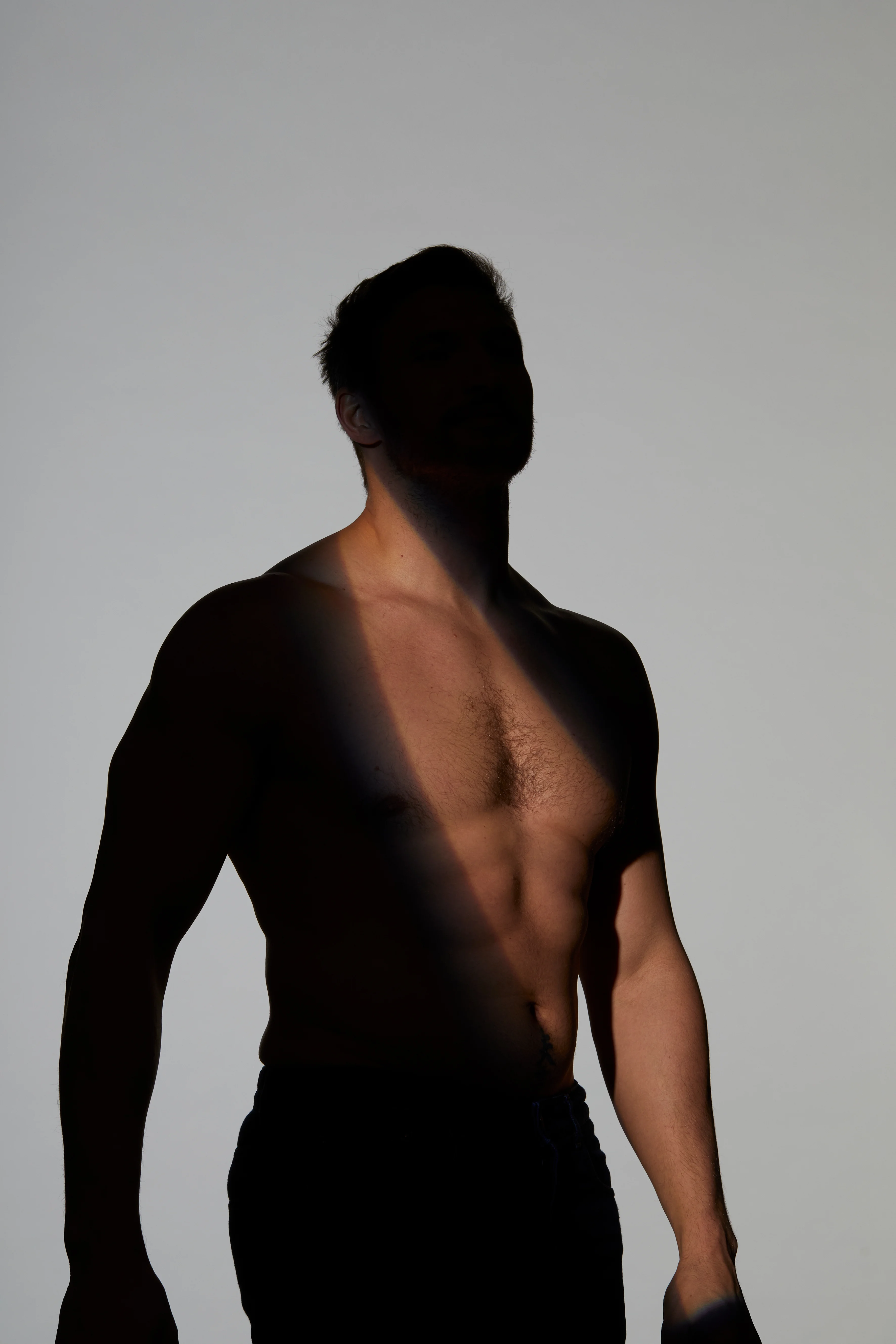 Ästhetische Aufnahme eines männlichen Oberkörpers im Halbschatten vor hellem Hintergrund, bei der das Licht die Brust- und Bauchmuskulatur betont – zur Veranschaulichung einer Brustverkleinerung beim Mann oder Gynäkomastie-Behandlung.